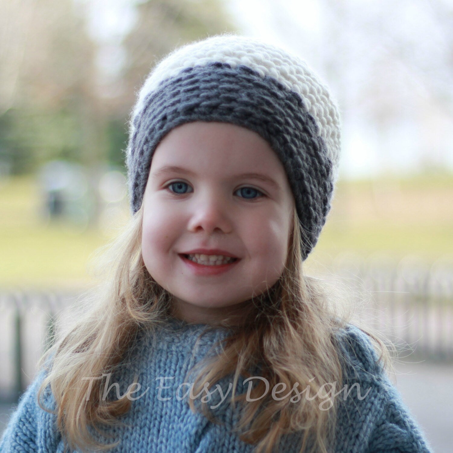 May include: A young girl with long blonde hair is wearing a gray and white crocheted hat. She is smiling and looking at the camera.