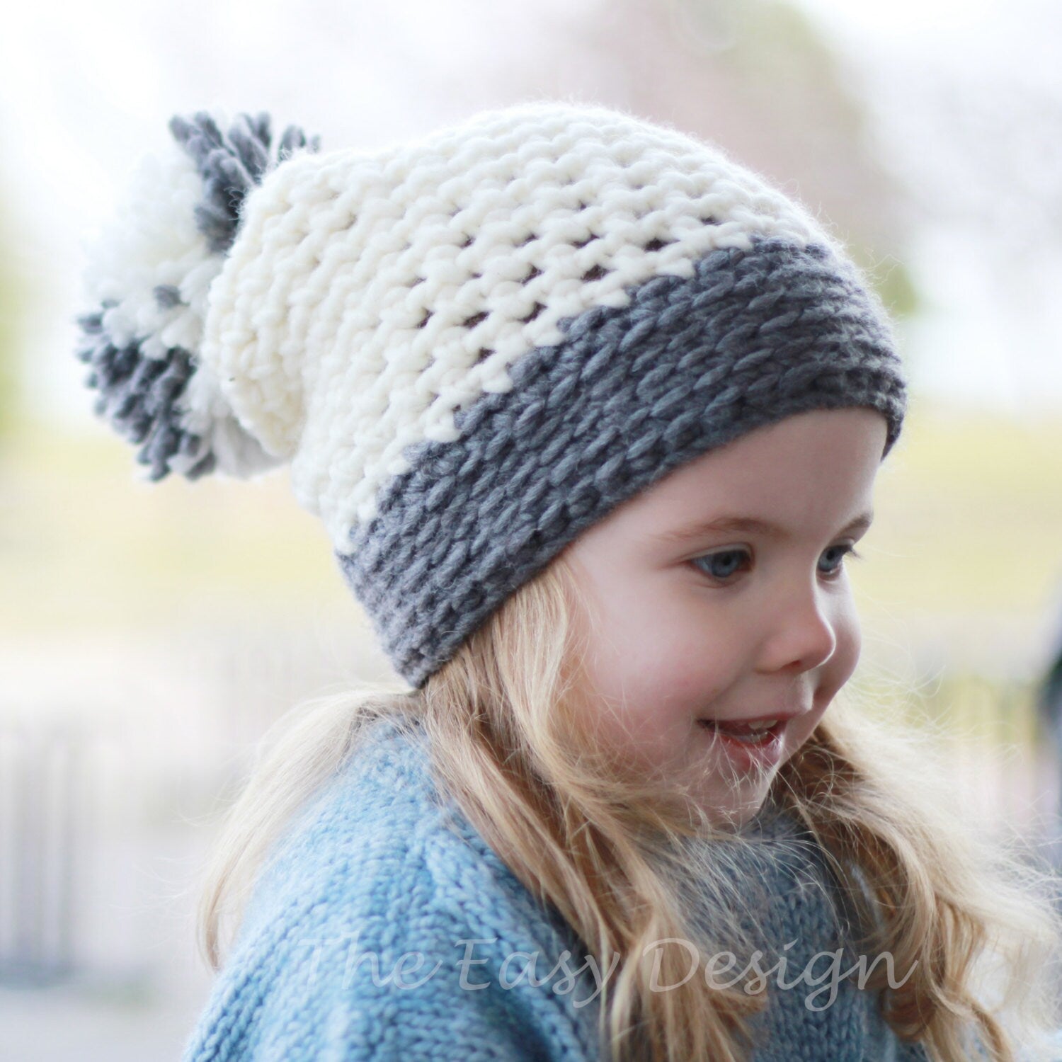 May include: A white and gray crocheted beanie with a fluffy pom pom on top. The beanie is worn by a child with long blonde hair.